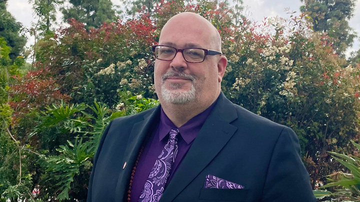A man in a purple tie stands in front of greenery.