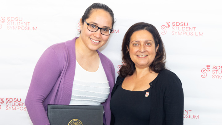 A woman in a purple cardigan holds an award while standing next to a woman in black.
